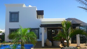 Un bâtiment moderne à deux étages, doté d’une piscine, de palmiers et avec une montagne en arrière-plan.