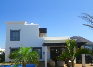 A modern two-story building with a pool, palm trees, and a mountain in the background.