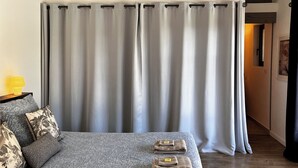Un dormitorio con cama, almohadas, una mesita de noche y un ventanal con cortinas.
