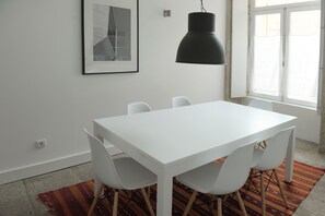 A white dining table with four chairs, a black pendant light, a framed picture on the wall, and a window with curtains.