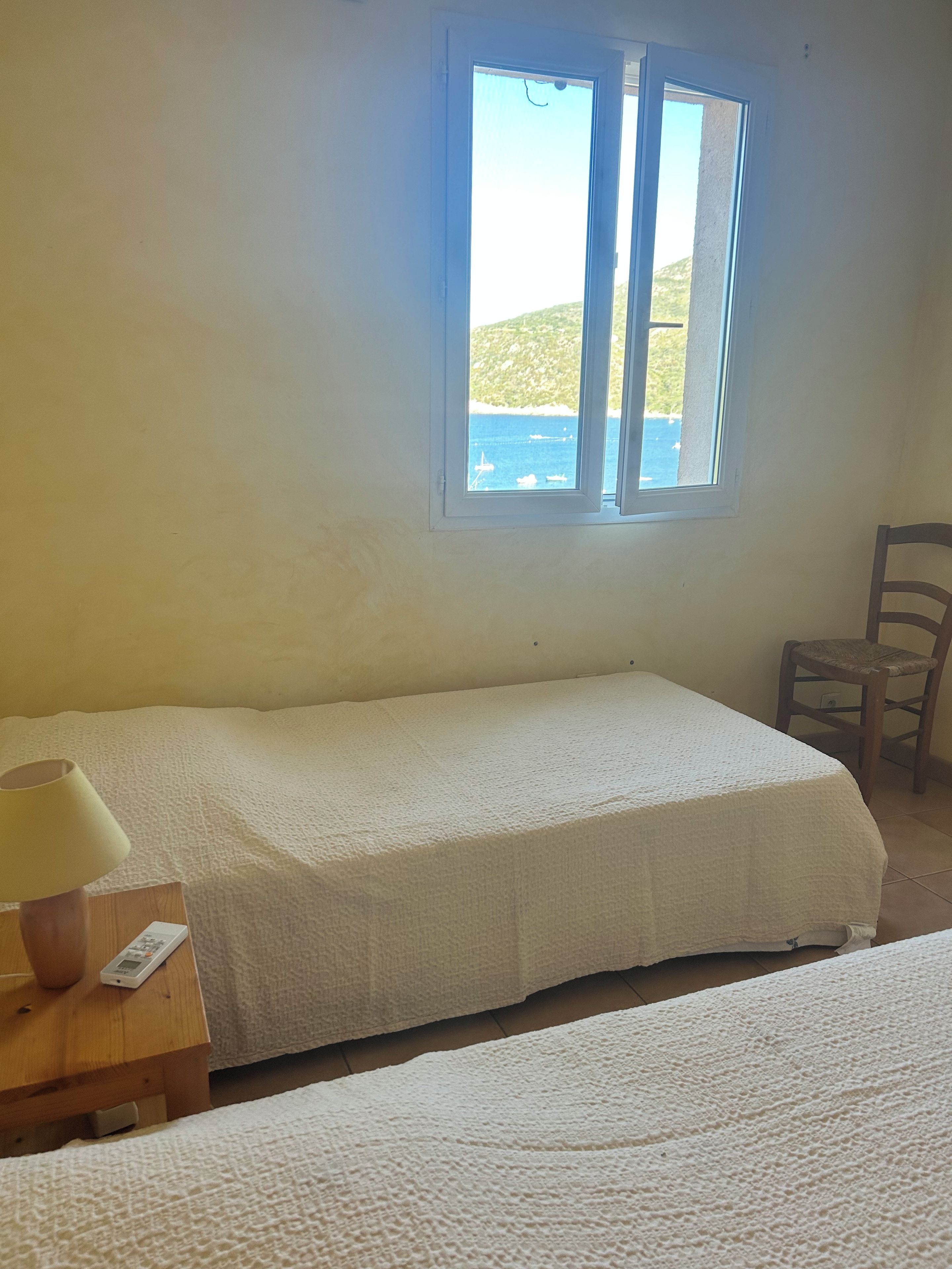 A single bed with a white bedspread, a bedside table with a lamp, and a window with a view of the sea.