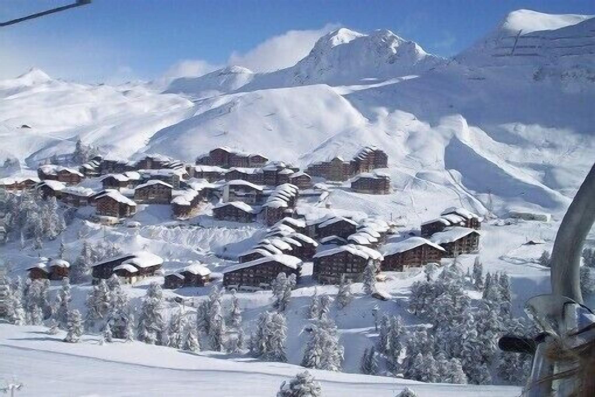 Un village de montagne enneigé, avec des chalets en bois, entouré d’arbres et de montagnes couverts de neige.