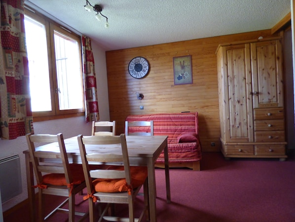 A room with a wooden floor, a wooden table with chairs, a red sofa, a wooden wardrobe, and a wall clock.