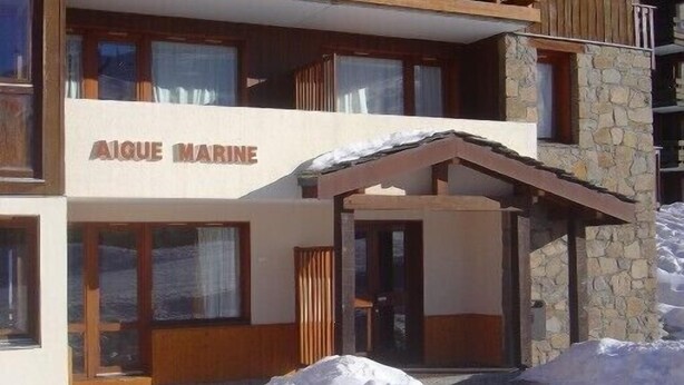 Ein zweigeschossiges Gebäude mit der Aufschrift 'AIGUE MARINE' auf der Fassade. Ein überdachter Eingang mit Holzboden und Steinwänden ist vorhanden.