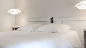 Cama com roupa de cama branca e duas almofadas brancas, cabeceira de madeira e duas luminárias pendentes.