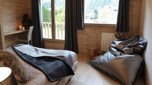 A wooden cabin room with a bed, a chair, a table with candles, and a view of a forested area through large windows. - 1/2 Chalet close to the center, quiet, private parking, 4 bedrooms 2 bathrooms (Chamonix-Mont-Blanc)