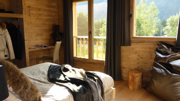 A wooden cabin bedroom with a bed, a chair, a small table, and a window overlooking a balcony. - 1/2 Chalet close to the center, quiet, private parking, 4 bedrooms 2 bathrooms (Chamonix-Mont-Blanc)