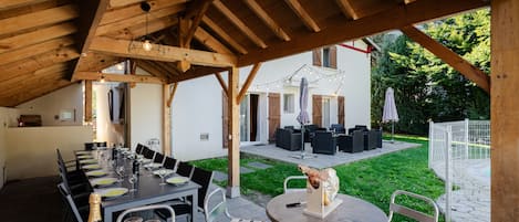 Un espace de restauration en plein air avec une longue table dressée pour un repas, entourée de chaises et surmontée d’une pergola en bois.