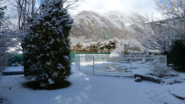 Een besneeerd landschap met een berg op de achtergrond, een omheind gebied met een bank en een met sneeuw beladen boom in de voorgrond.