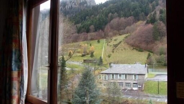 A room with a large window offering a view of a mountainous landscape, a building, and a parking area. - Residence A. LE CAPUCIN, N ° 68 Apartment 4 people facing LUGE TENNIS COURTS (Mont-Dore)