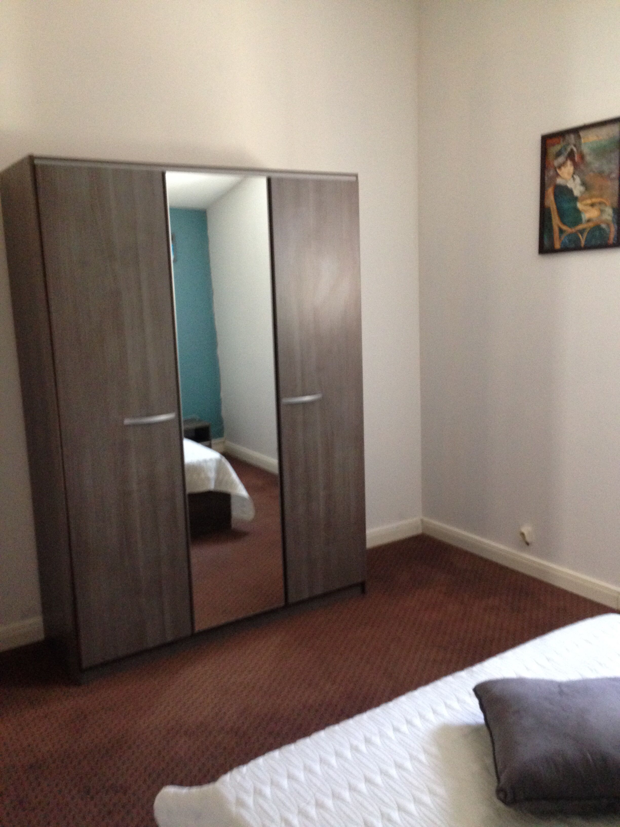 A bedroom with a large wardrobe, a bed with a white comforter, and a painting on the wall.