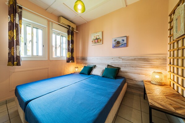 A bedroom with a large bed, two windows with curtains, a wooden headboard, and a small table with a lamp.