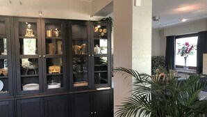 Un salón moderno con un mueble de madera oscura que contiene objetos decorativos y cristalería. Hay una planta grande en primer plano.