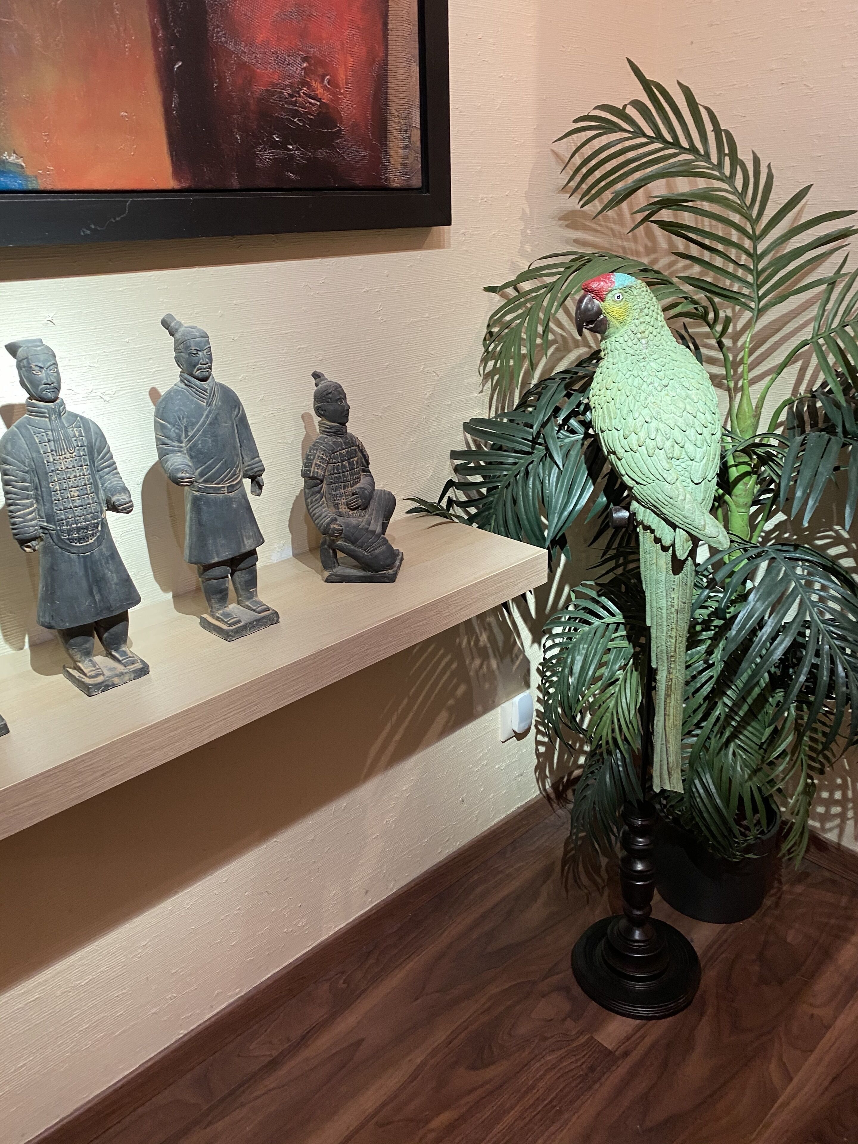 Ein Regal mit drei Terrakotta-Statuen, einem Papagei auf einer Pflanze und einem Gemälde an der Wand.