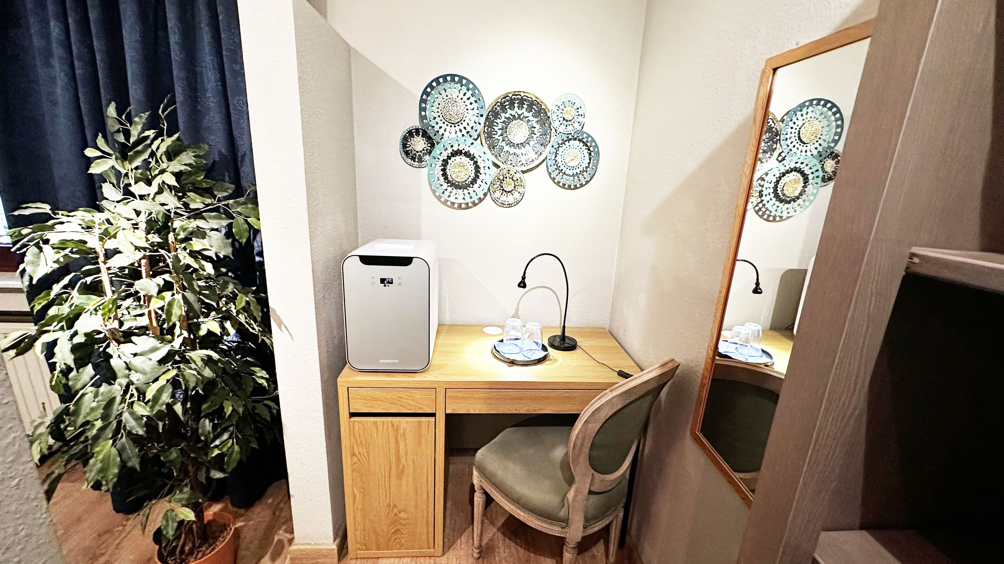 A compact room with a desk, chair, and a large mirror. A small plant is placed beside the desk, and decorative wall art is visible above the mirror.