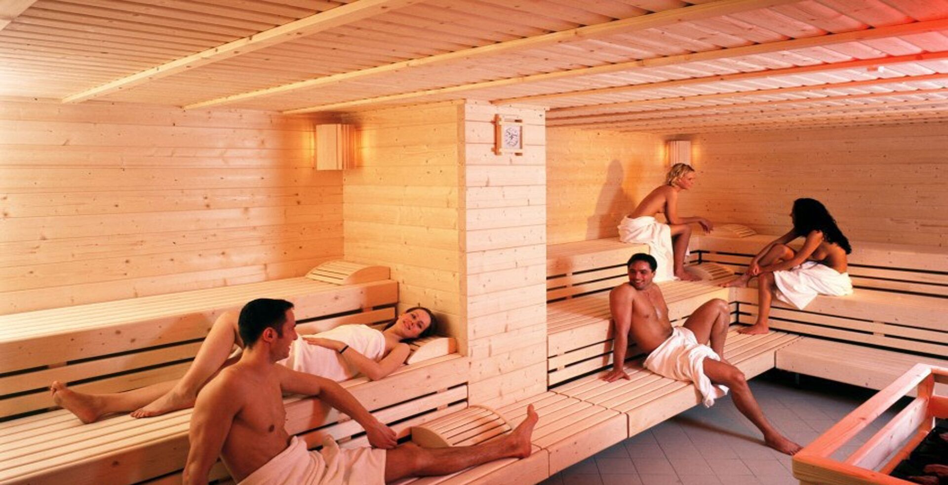 Eine Sauna mit Holzbänken, einer sichtbaren Uhr und einer sitzenden Person auf einer Bank.