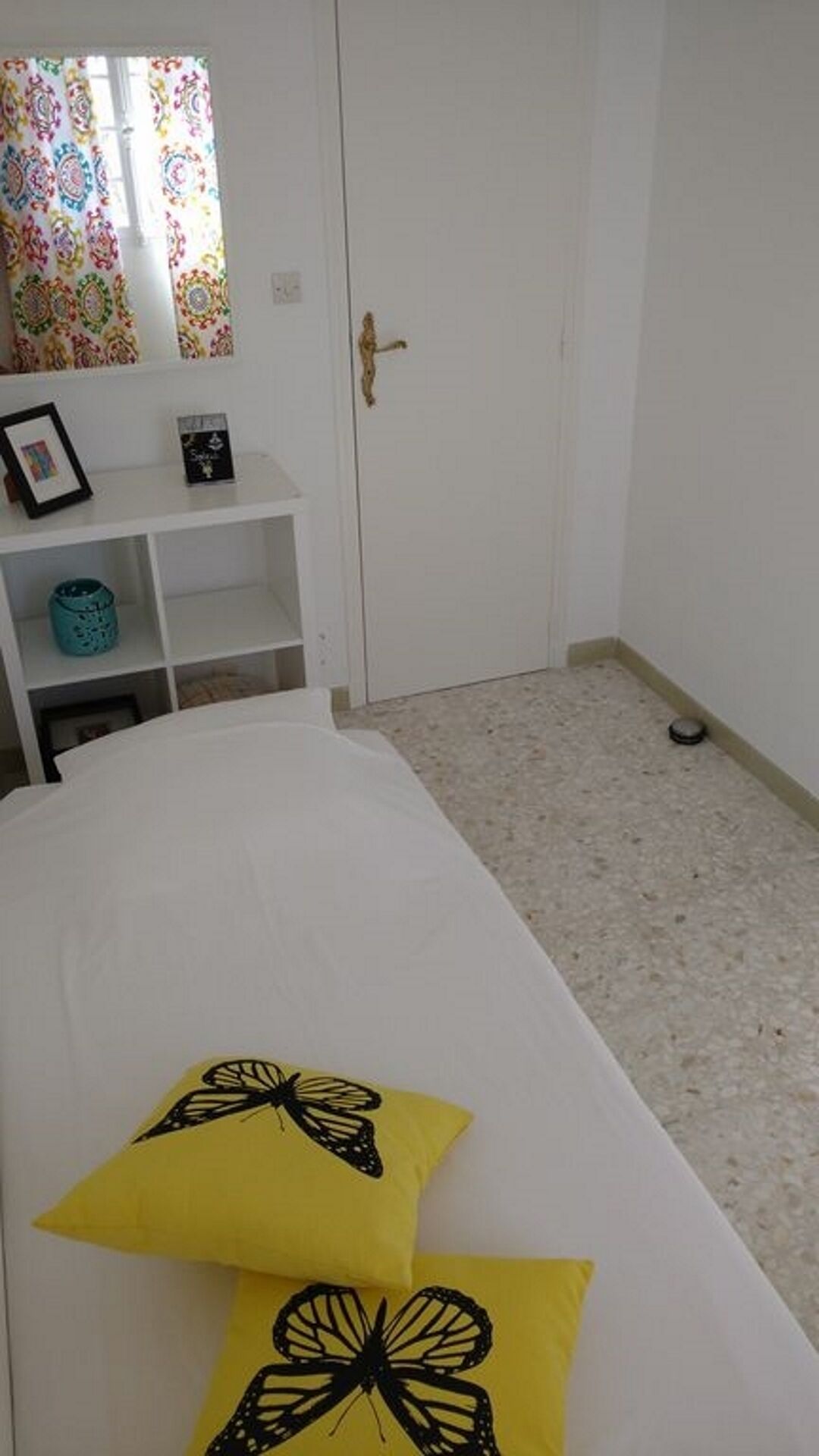 Un dormitorio con cama, dos almohadas amarillas con diseños de mariposas negras, un armario blanco, una estantería con un cuadro enmarcado y un libro, y una cortina colorida.