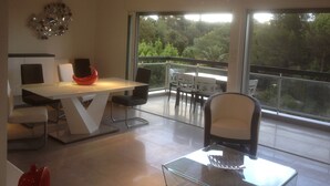 A modern dining area with a white table, black chairs, and a red bowl. There is a balcony with a view of trees. - Charming apartment in property with pool and large park (Saint-Raphaël)