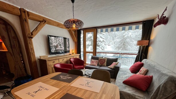 Een gezellige kamer met een houten eettafel, rode stoelen, een grijze bank met rode kussens en een televisie aan de muur.