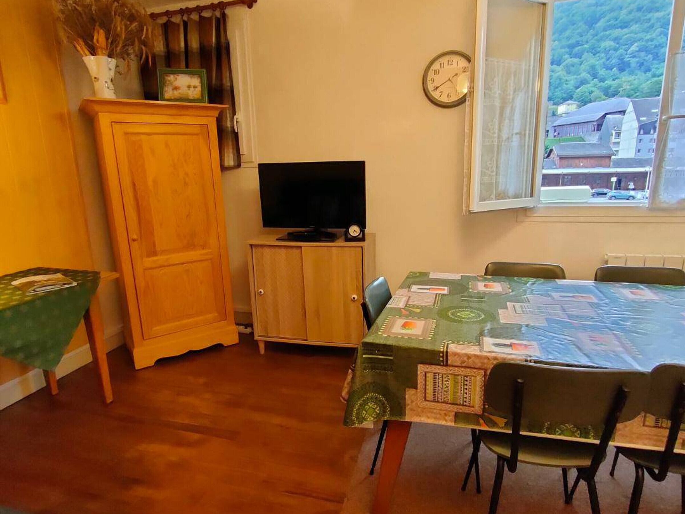 A room with a wooden cabinet, a television on a stand, a dining table with chairs, and a clock on the wall.