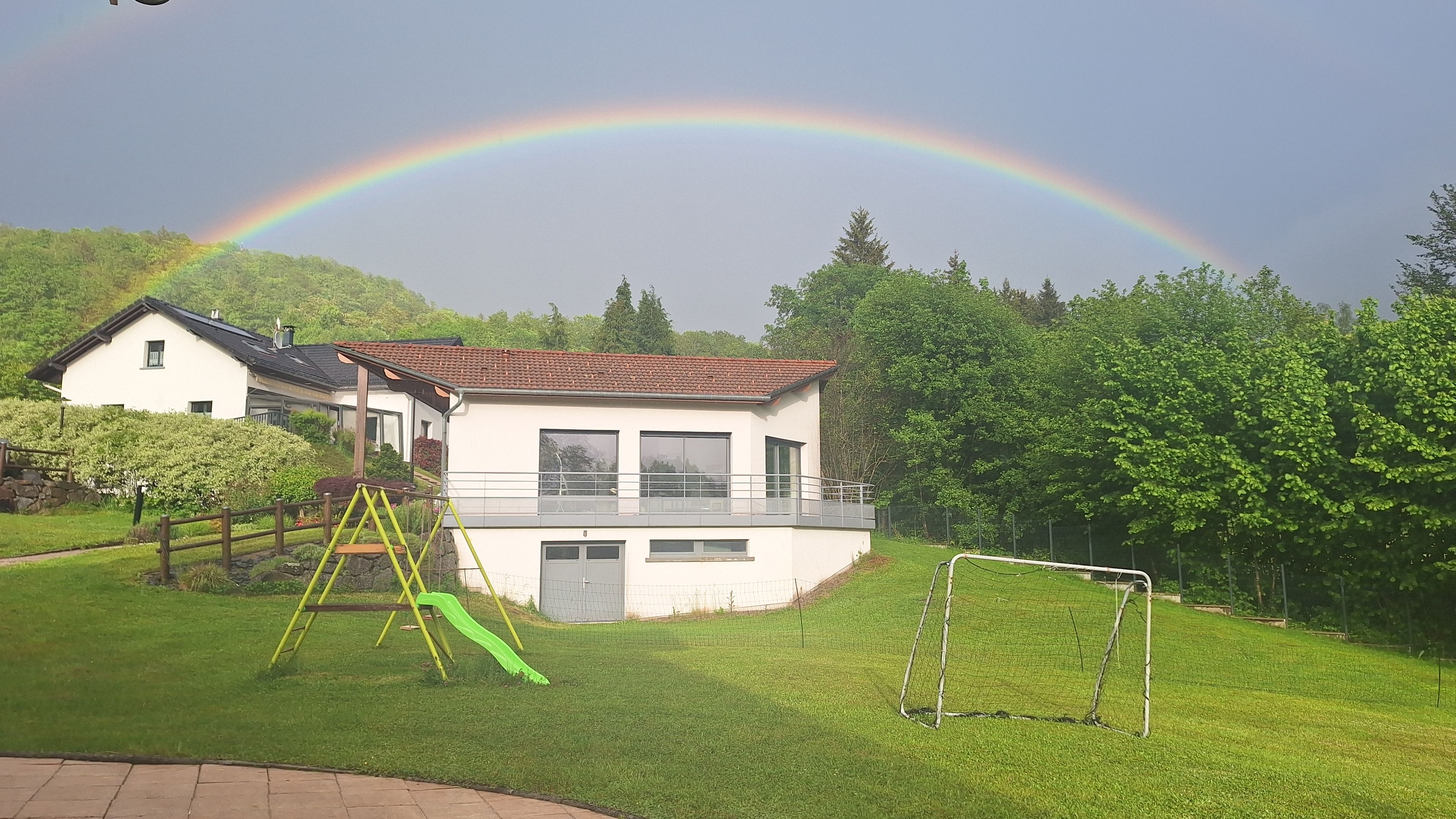 Une maison avec un balcon, un toboggan vert et un but de football dans une cour gazonnée.