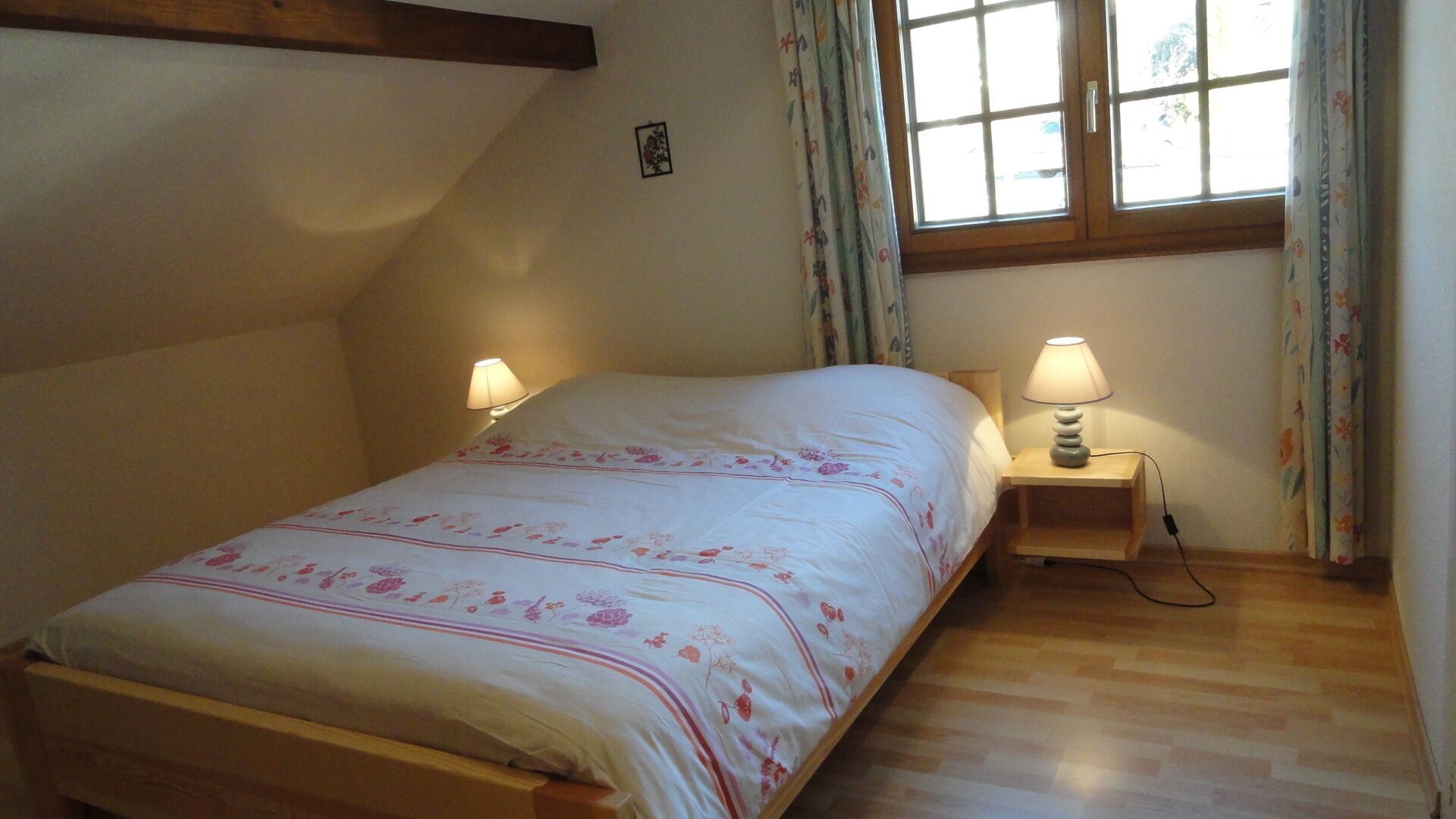 Une chambre à coucher avec un lit, deux lampes de chevet, une fenêtre avec des rideaux et un plancher en bois.