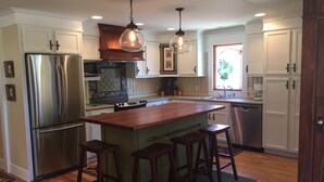 Una cocina con gabinetes blancos, electrodomésticos de acero inoxidable, una isla de madera con taburetes, iluminación colgante y una ventana con vistas a la vegetación.