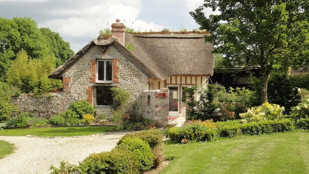 Ein traditionelles Steinhaus mit einem Reetdach, umgeben von einem gepflegten Garten und einem Kiesweg, der zum Eingang führt.