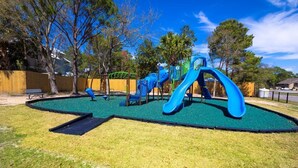 A playground with slides and climbing structures, surrounded by grass and trees.