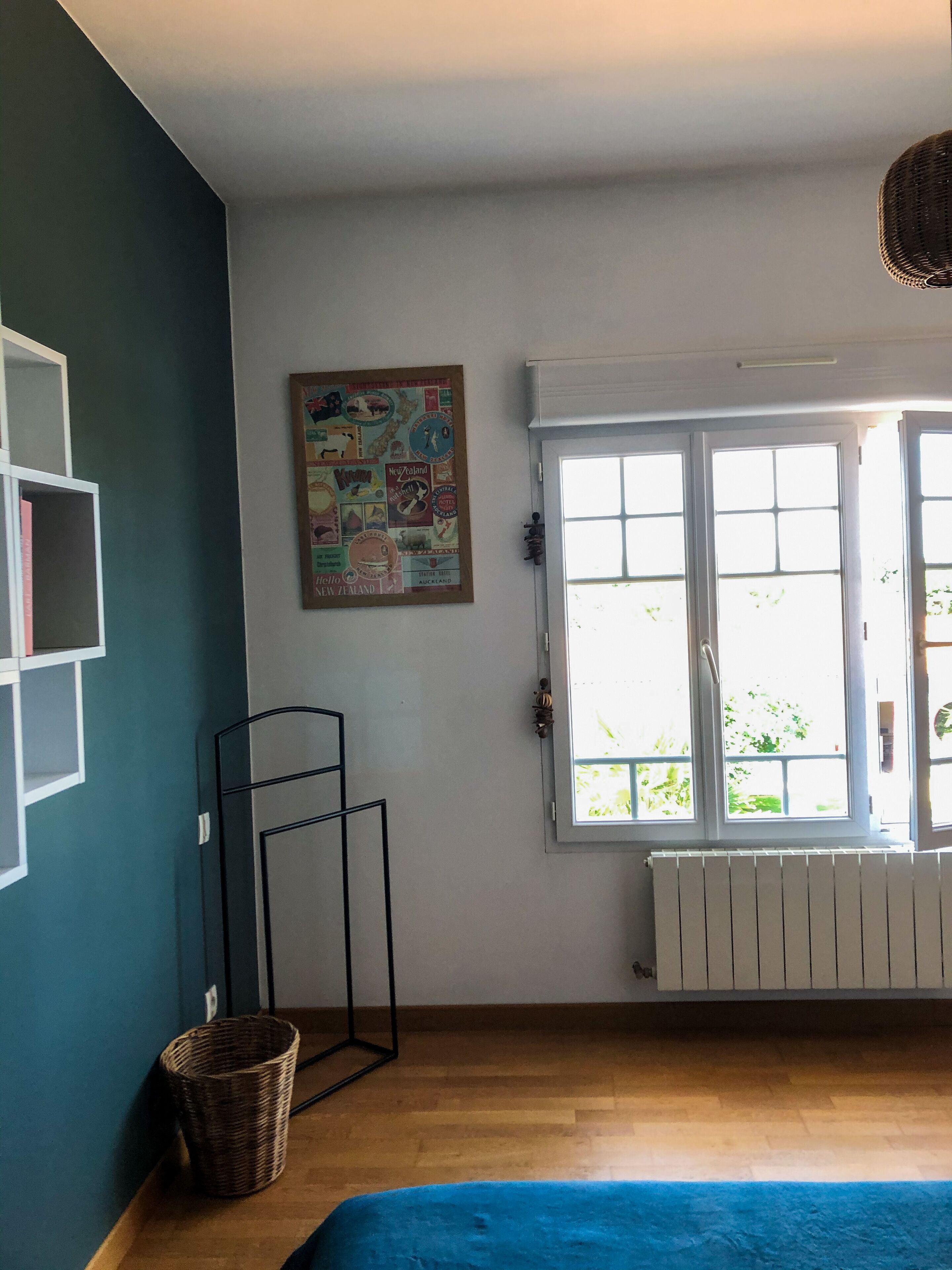 A room with a large window, a radiator, a blue sofa, a wicker basket, and a wall with a travel-themed poster.