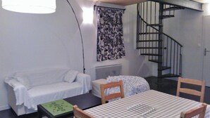 Una habitación con un sofá blanco, una mesita con un libro verde, un mantel a cuadros y una escalera de caracol.