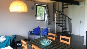 Un comedor con una mesa dispuesta para cuatro personas, un sofá con cojines azules, una escalera y un ventanal con cortinas.