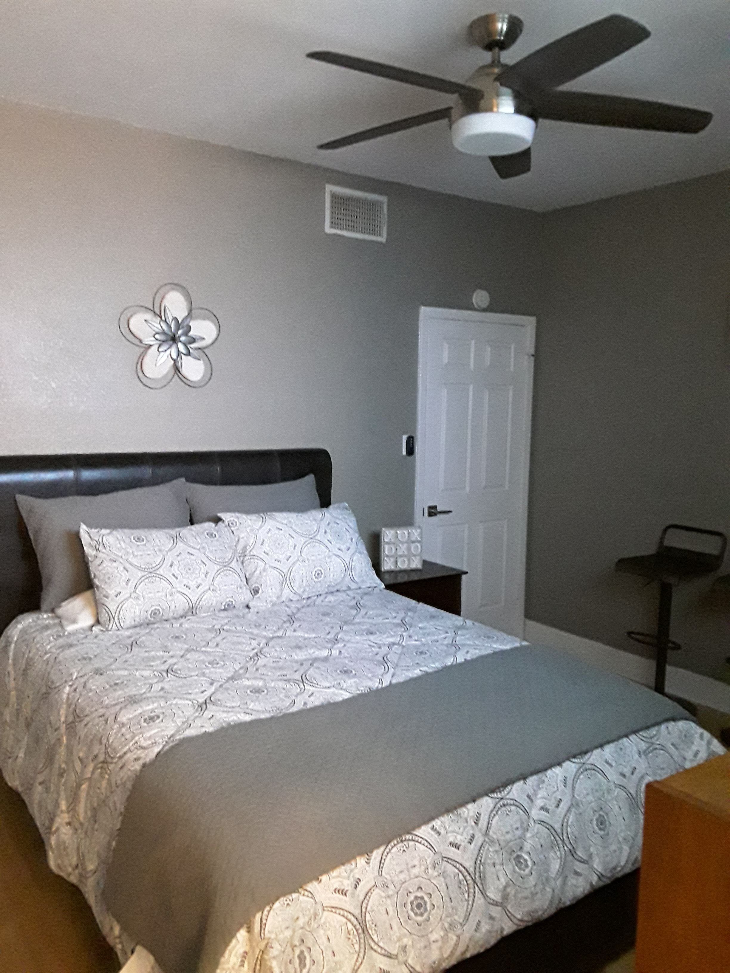 A bedroom with a bed, a ceiling fan, a door, and a small table.