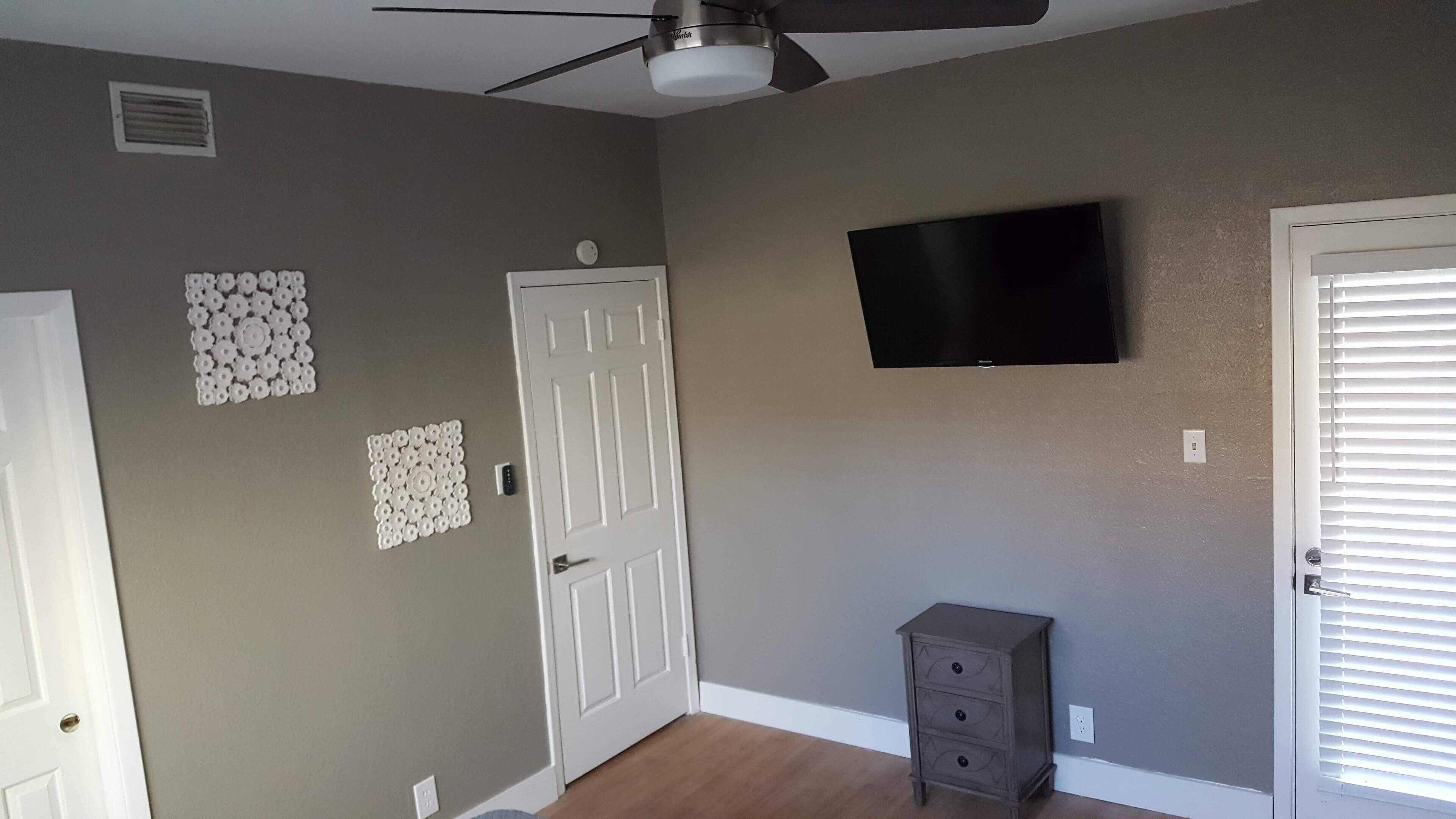 A room with a ceiling fan, a flat-screen TV mounted on the wall, and a small cabinet against the wall.
