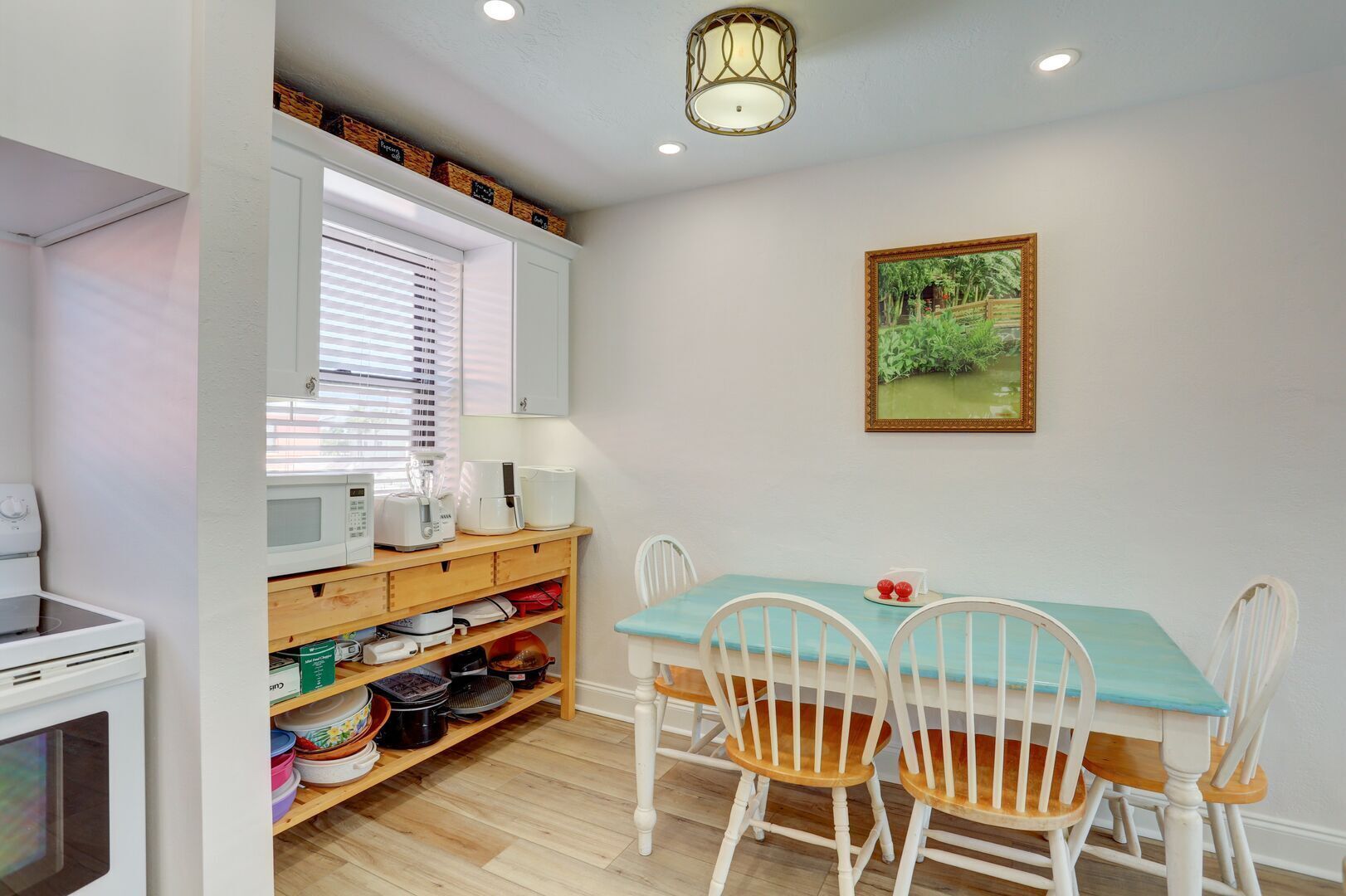 Une cuisine compacte avec un coin repas, comprenant une table en bois, des chaises blanches et une fenêtre avec des stores.