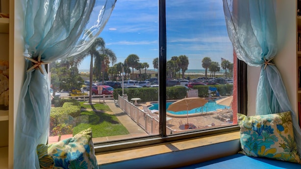 A room with a large window overlooking a pool area, palm trees, and a parking lot. - Newly Remodeled Poolside Condo with Gorgeous Ocean-Front View - Sleeps 8 (St Pete Beach)