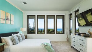 A bedroom with a large bed, a dresser, a TV, and a view of the ocean through multiple windows.