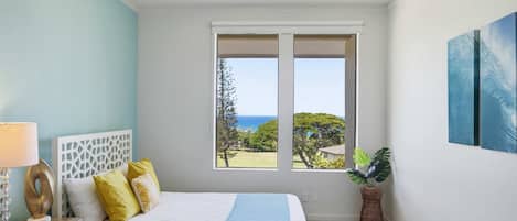 Un dormitorio con cama, mesita de noche, lámpara, ventana con vista al océano y una planta en maceta.