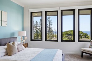 Un dormitorio con una cama grande, una mesita de noche, una lámpara y vistas a árboles a través de tres ventanas.