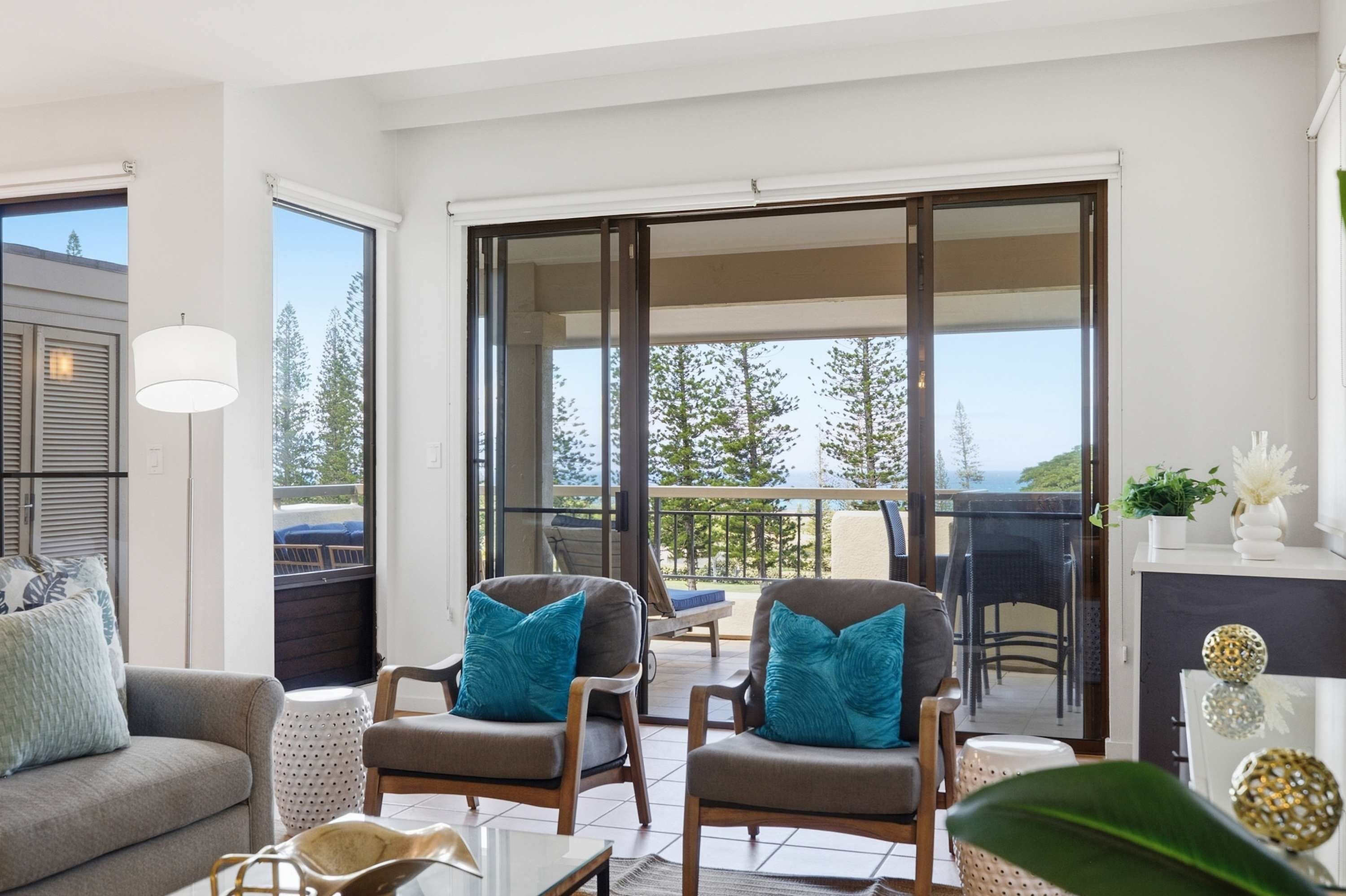 A modern living room with a sofa, two armchairs, a coffee table, and a balcony with a view of trees and the sky.