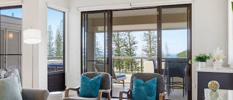 Una sala de estar moderna con un sofá, dos sillones, una mesa de centro y un balcón con vistas a árboles y al cielo.