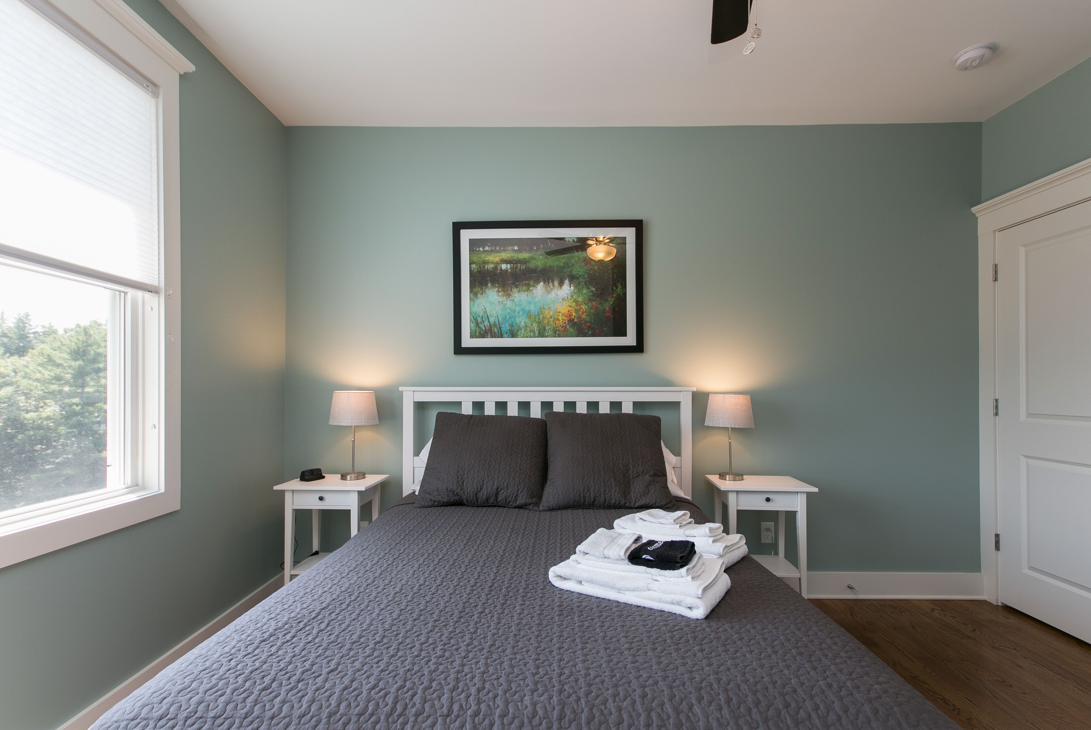 A neatly made bed with a grey quilt, two bedside lamps, a framed picture of a landscape, and a window with blinds.