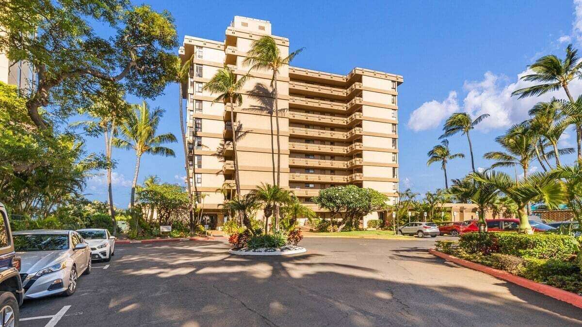 Un hotel de varios pisos con un patio central, rodeado de palmeras y un estacionamiento.