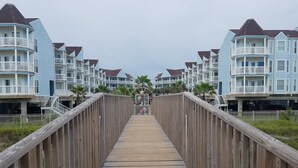 Un paseo marítimo de madera que conduce a una hilera de edificios azul claro con balcones y un arco central.