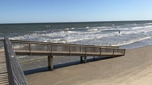 Un paseo marítimo de madera que lleva a una playa arenosa con el océano al fondo.