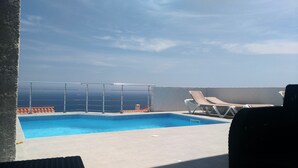 Una piscina al terrat amb vista a l'oceà, una barana de vidre i dues gandules.