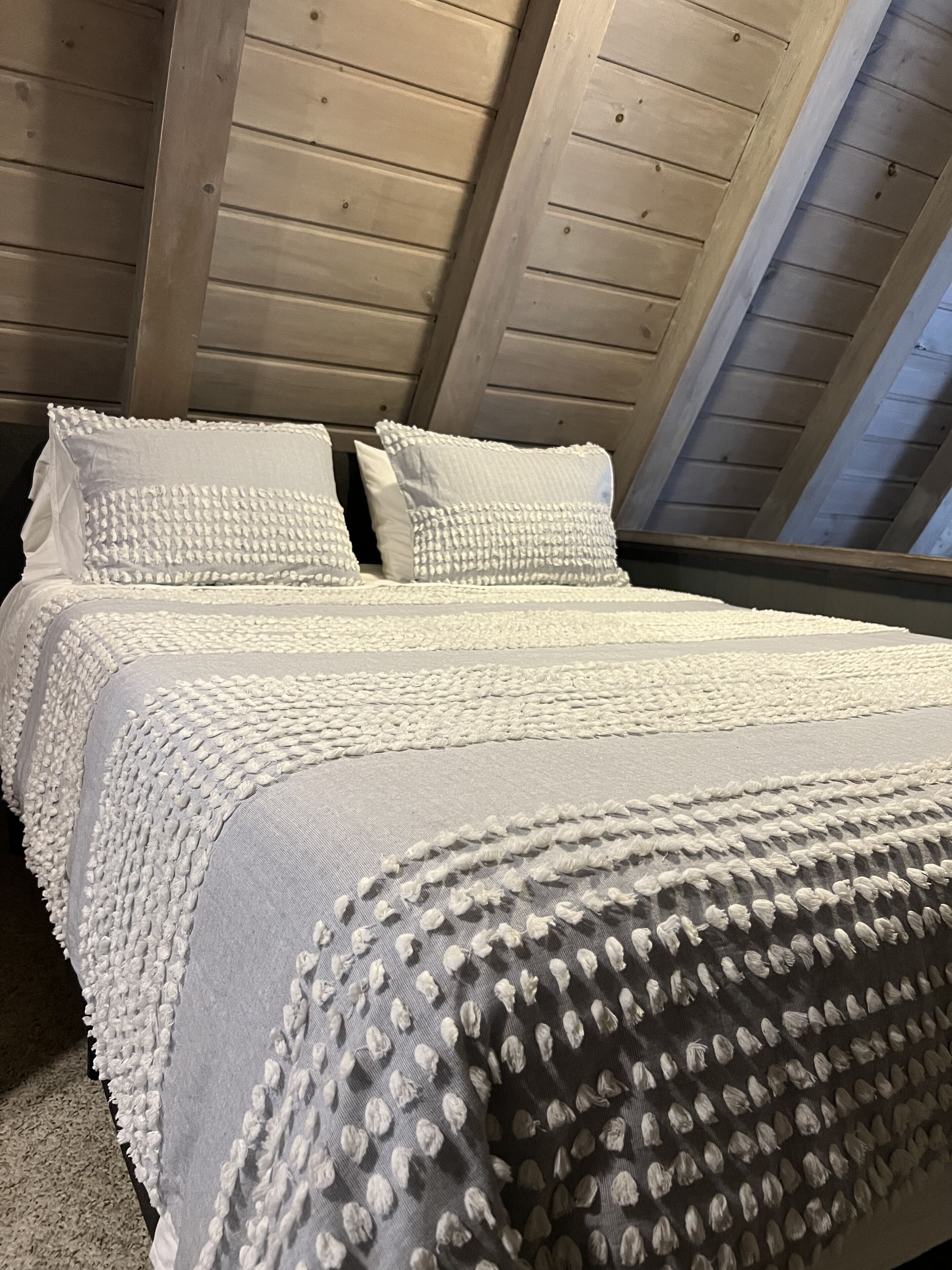 A bedroom with a wooden ceiling, a bed with white bedding, and a window with a view of the sky.