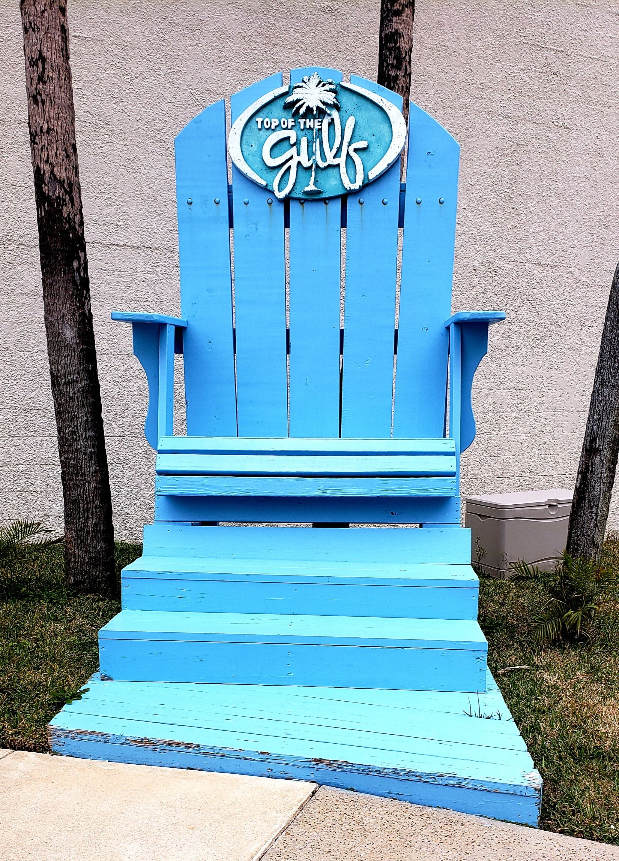 Une chaise Adirondack bleue, sur laquelle est fixée une plaque portant l’inscription « Au sommet du golfe ».