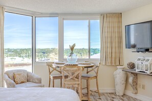 A hotel room with a large window, a dining table with chairs, a bed, a television, and a decorative vase. - Beachfront Studio, Free Beach Service included with stay (Panama City Beach)