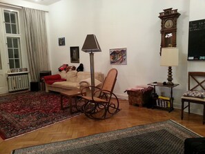 A living room with a sofa, rocking chair, and a small table with a lamp. There is a clock on the wall and a radiator near the window.